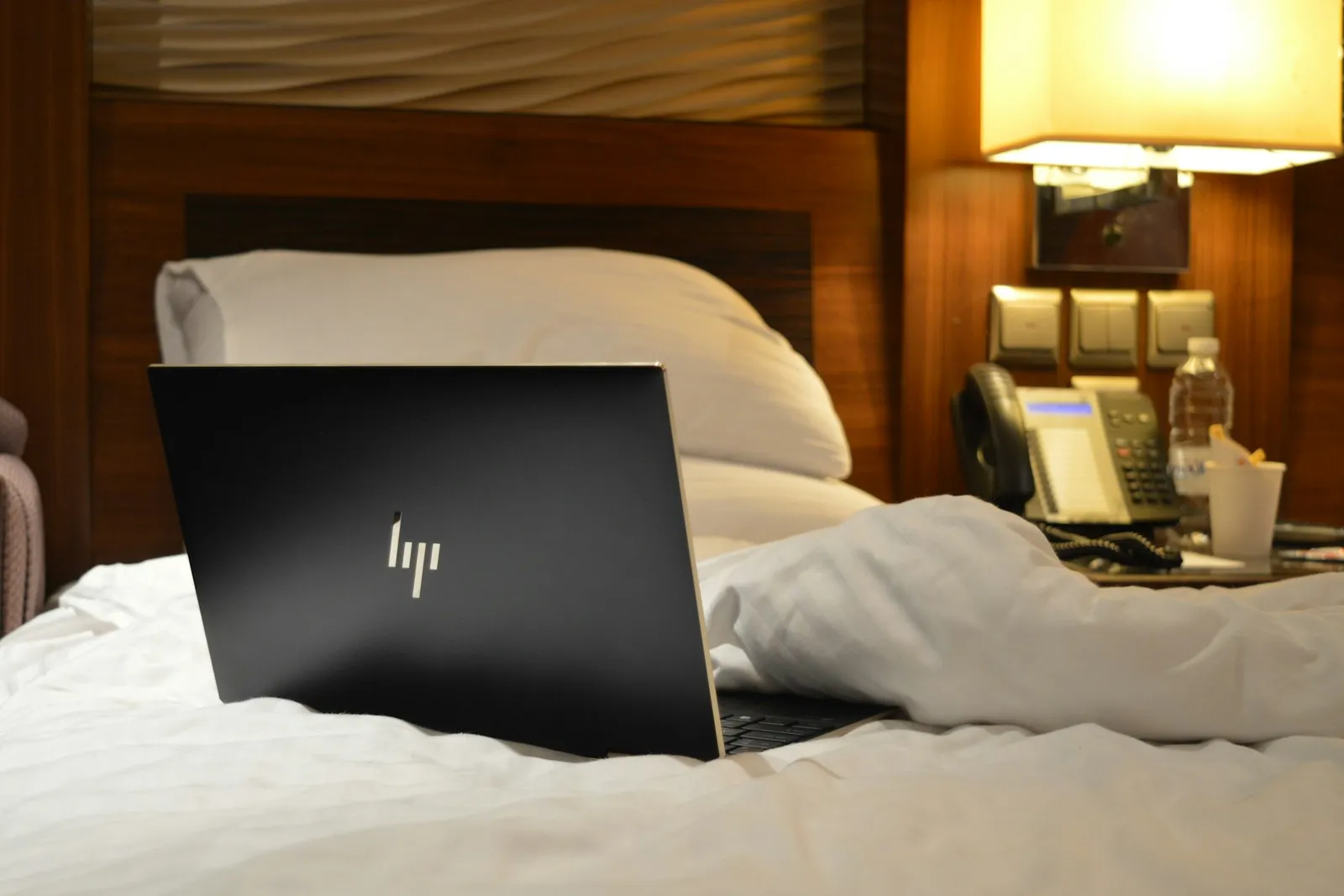 Laptop auf einem Hotelbett neben Nachttischlampe und Telefon